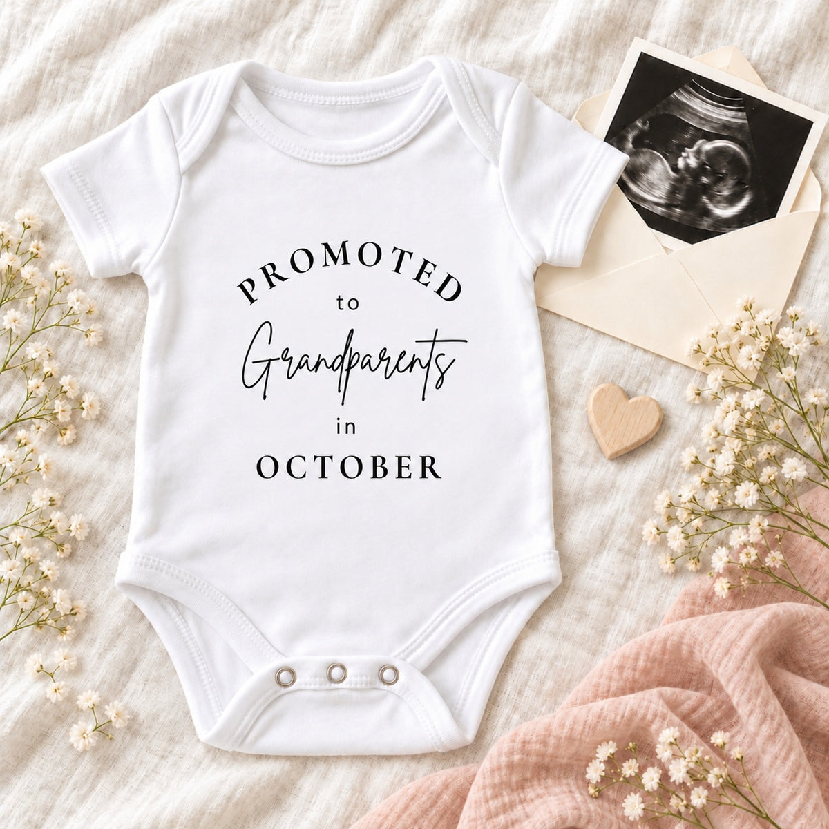 White Baby Vest printed with Promoted to Grandparents in October, styled with a scan photo, ivory envelope, wooden heart, blush muslin and delicate white flowers.