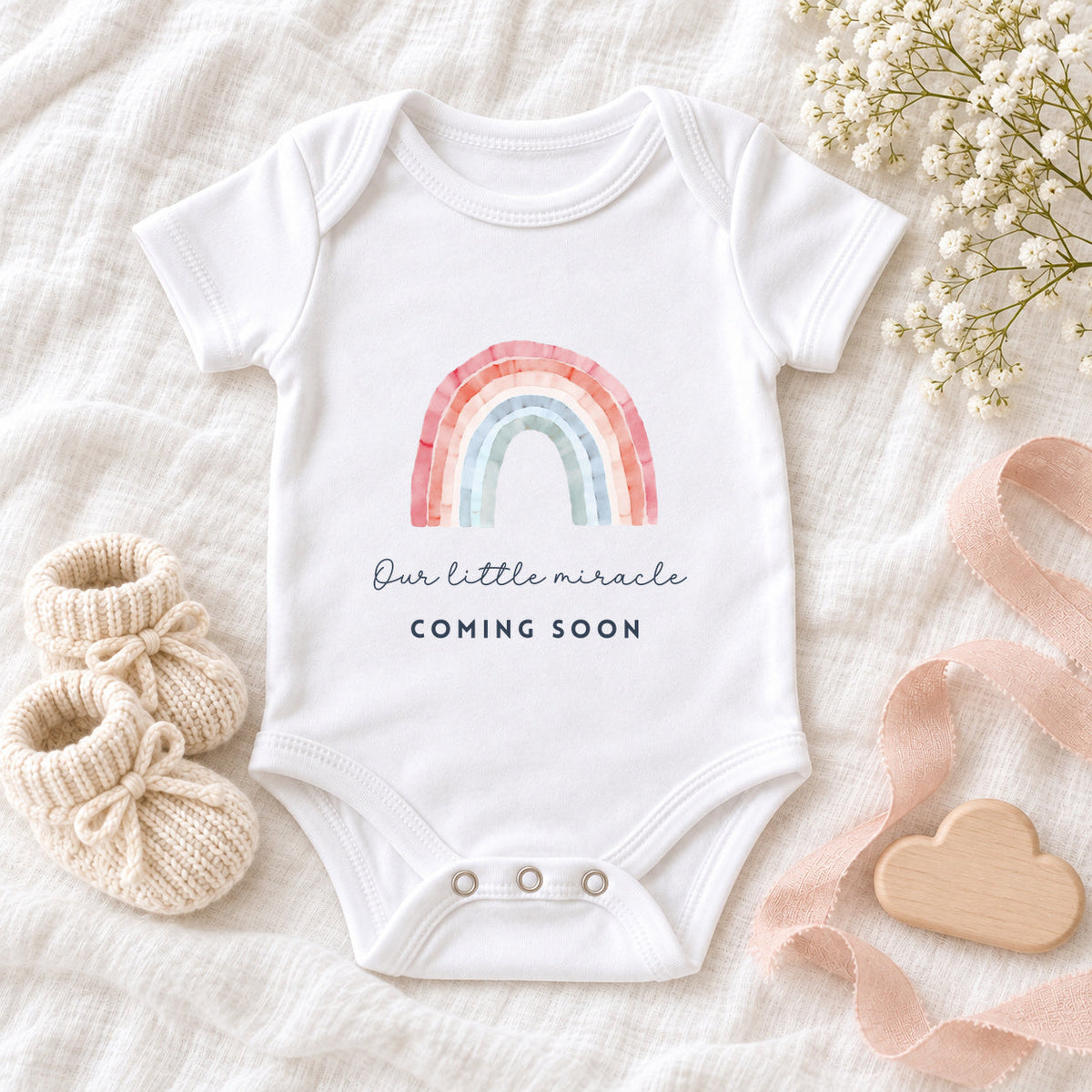 White Baby Vest with a pink, peach, blue and sage watercolour rainbow above 'Our little miracle' and 'COMING SOON', on white muslin with cream booties, a wooden cloud and blush ribbon.