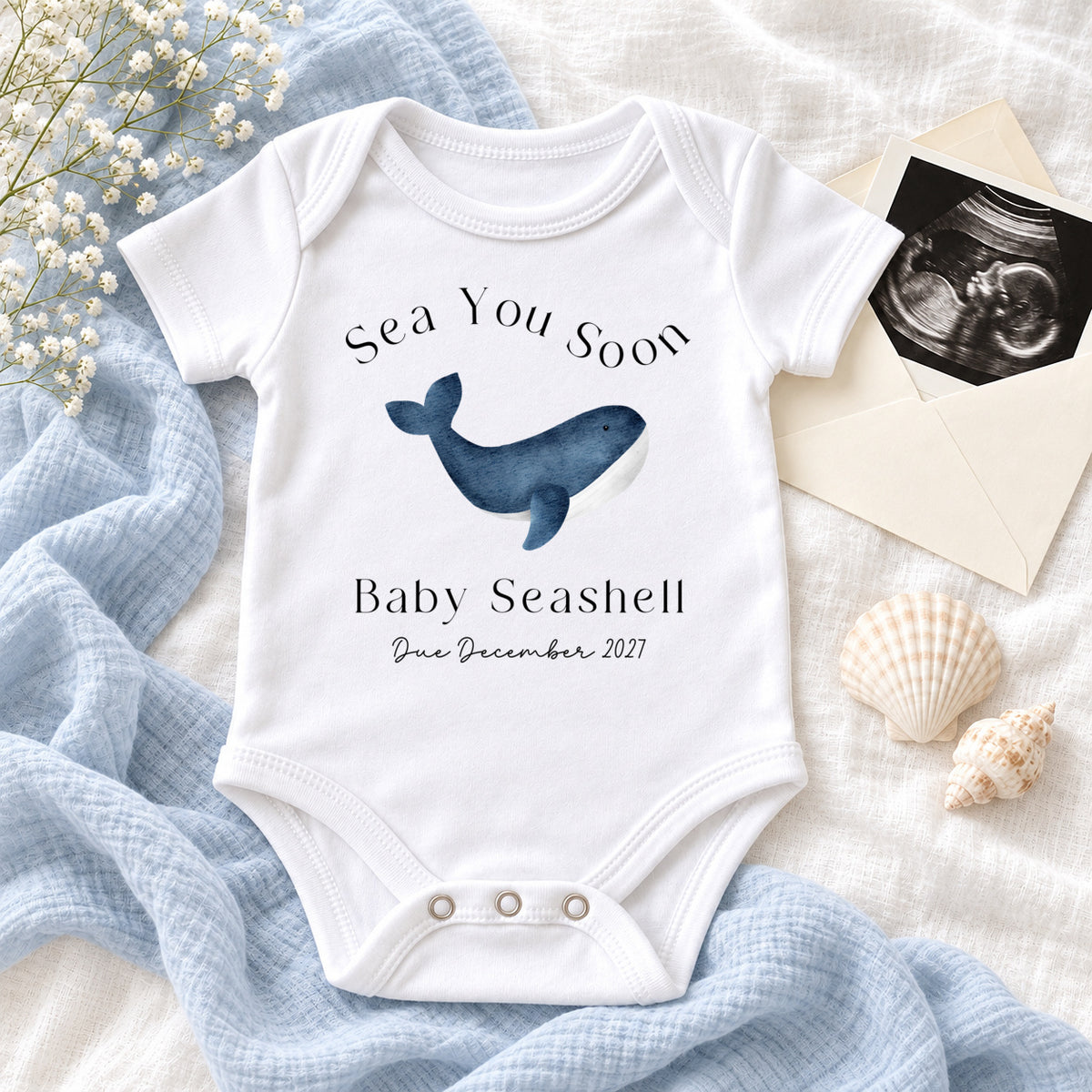 White Baby Vest printed with Sea You Soon, a blue watercolour whale, Baby Seashell and Due December 2027, styled with ivory knitwear, blush muslin, seashells, white flowers and a wooden whale.