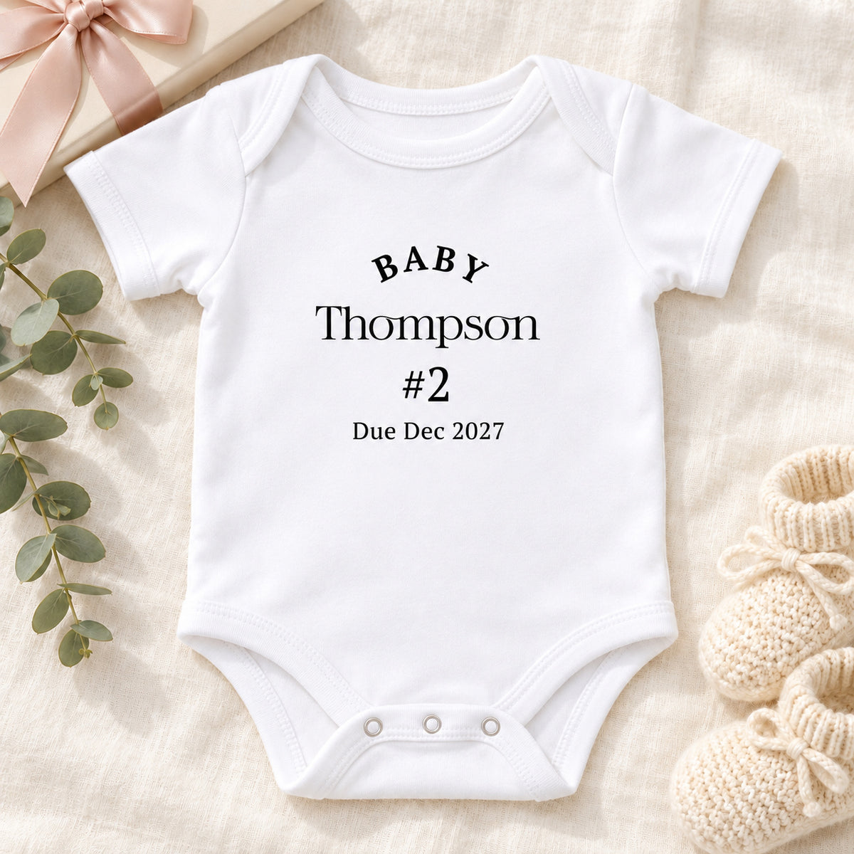 Second pregnancy announcement white Baby Vest with BABY Thompson #2 Due Dec 2027 print, cream gift box, ribbon, knitted baby booties and eucalyptus on beige linen, with props notice.