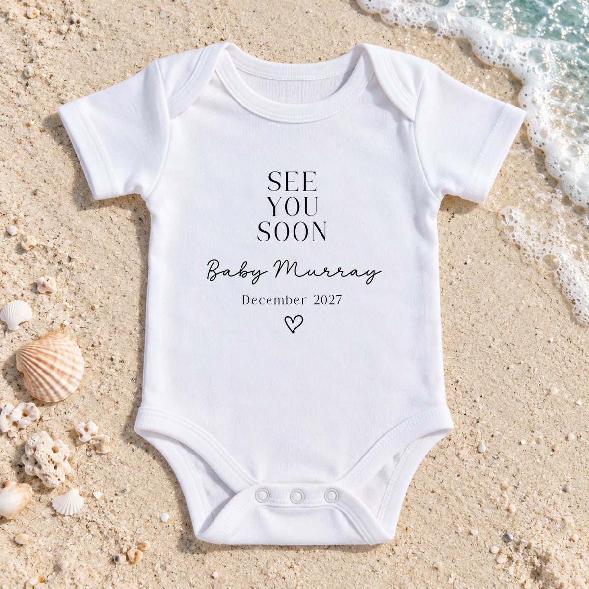 White See You Soon Baby Vest with Baby Murray, December 2027 and a heart, laid on pale beach sand beside seashells and turquoise sea foam.