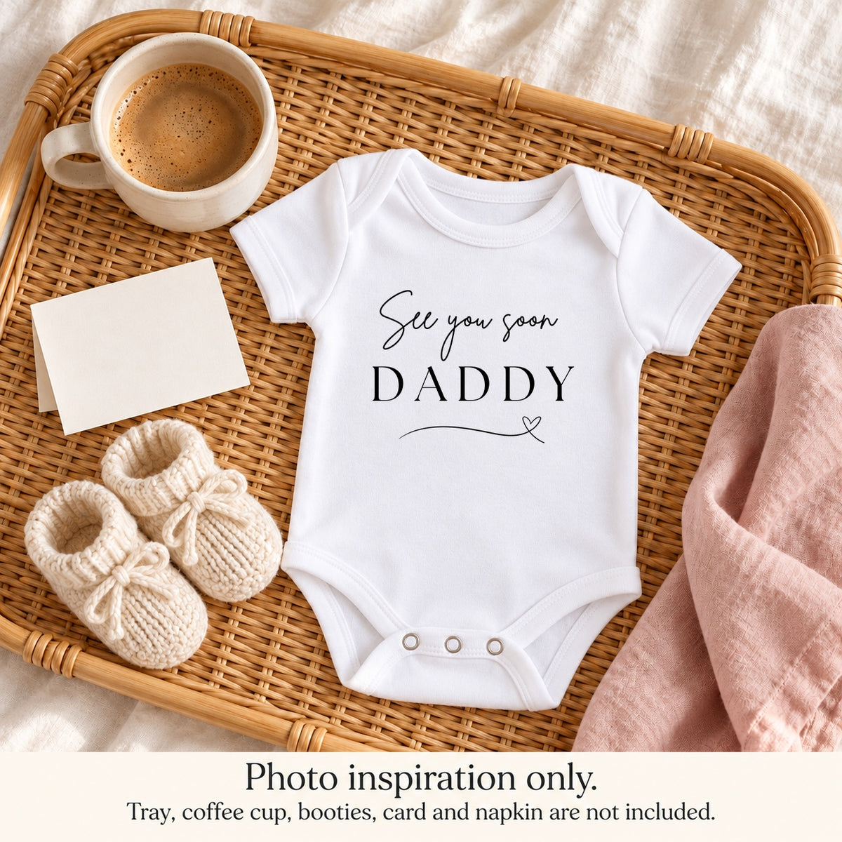 White See You Soon Daddy pregnancy announcement baby vest with a heart flourish on a rattan breakfast tray with coffee, cream booties, a blank card and blush napkin.