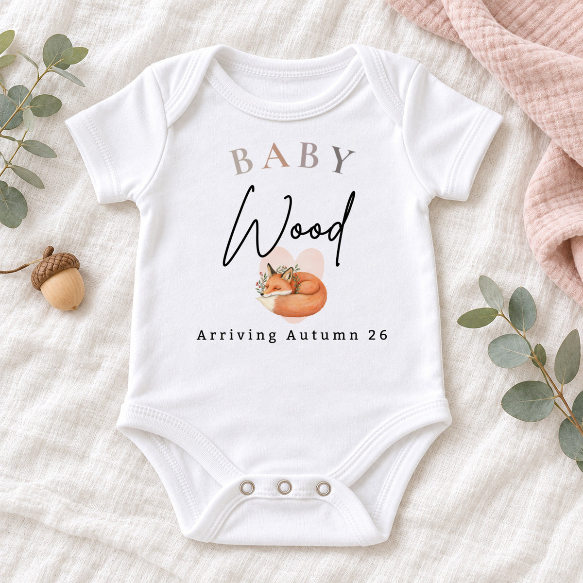 Personalised white Baby Vest with Baby Wood, sleeping fox and Arriving Autumn 26 artwork, styled with eucalyptus, an acorn and blush muslin