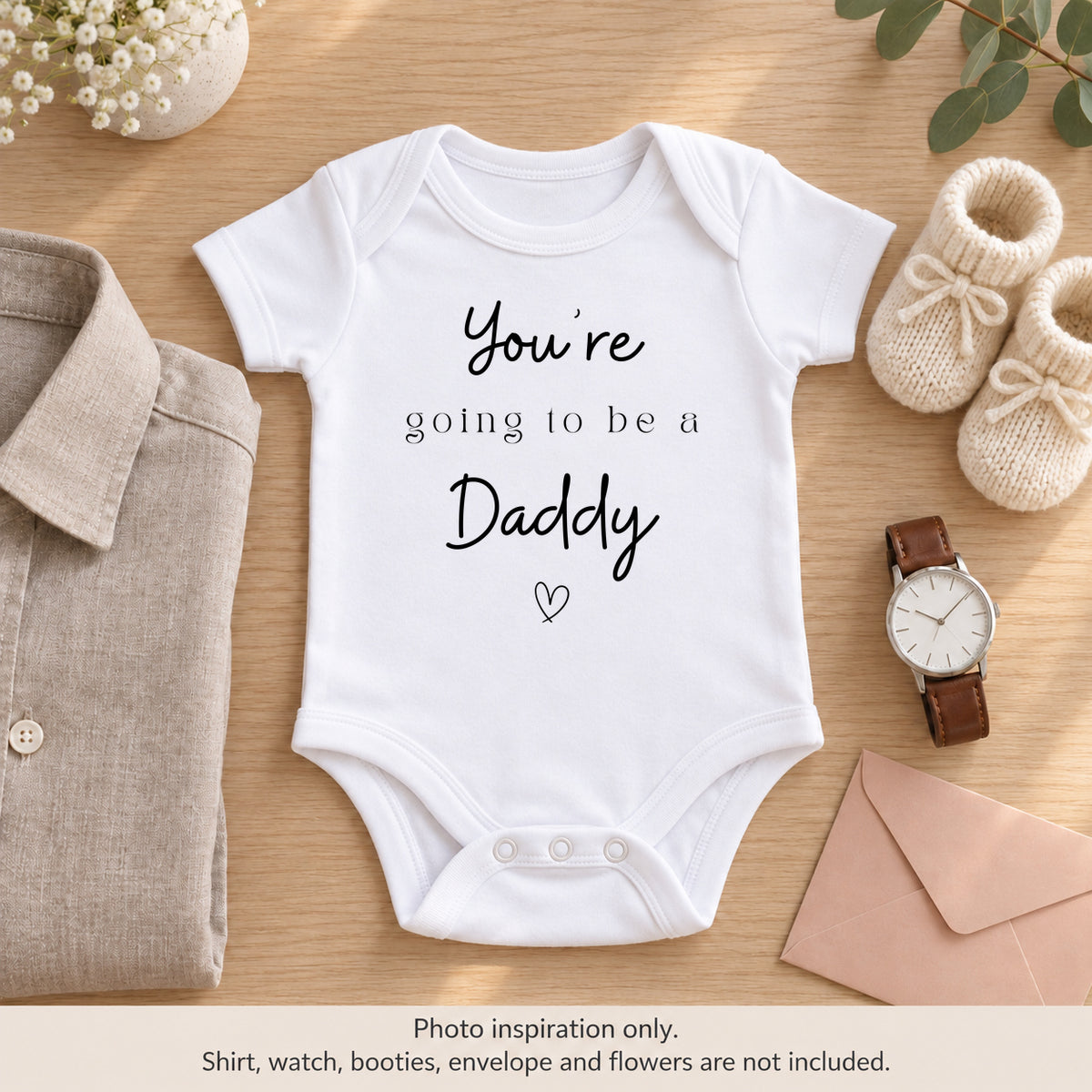 White 'You're Going to Be a Daddy' pregnancy announcement baby vest beside a neutral shirt, watch, cream booties and blush envelope.